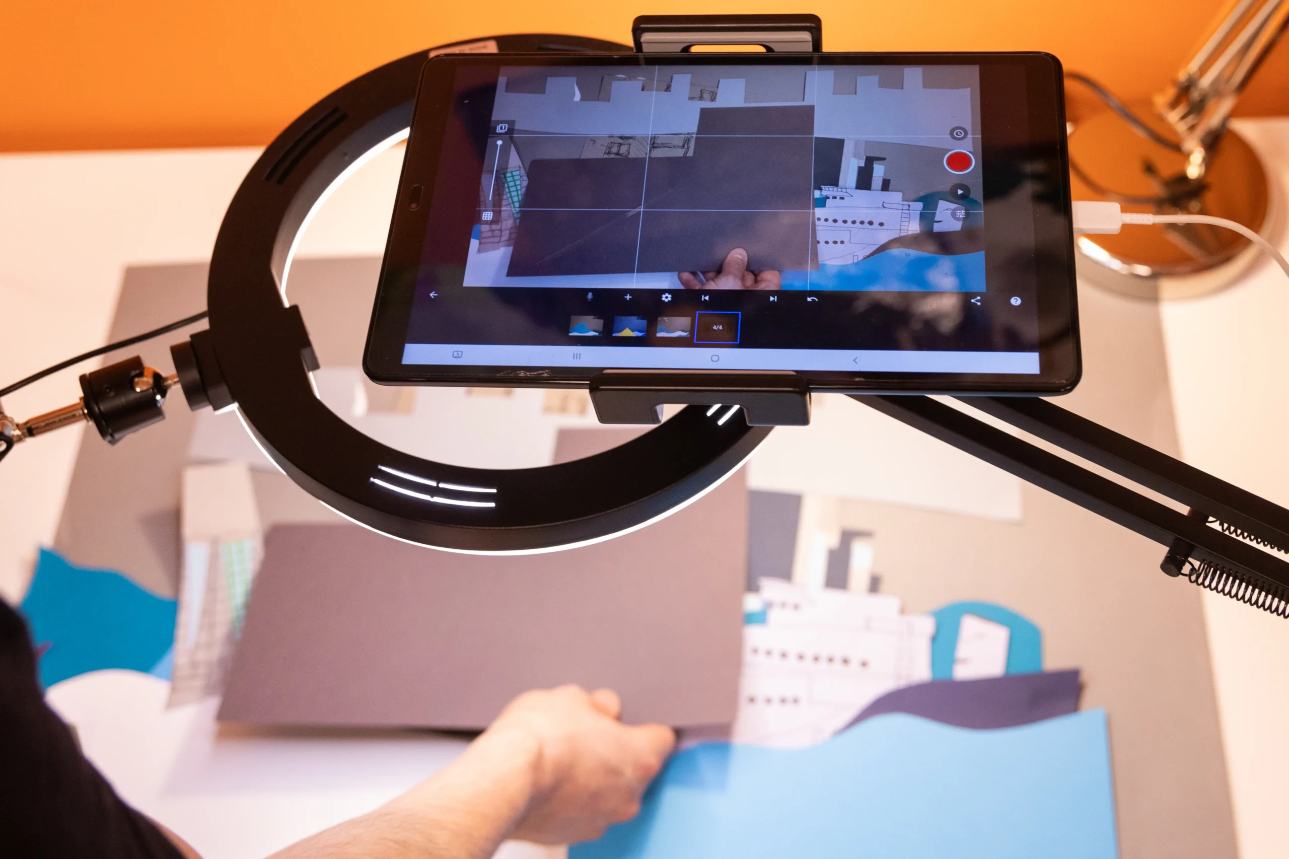 Dłoń układa papierowe elementy pod tabletem na statywie podczas tworzenia animacji poklatkowej.