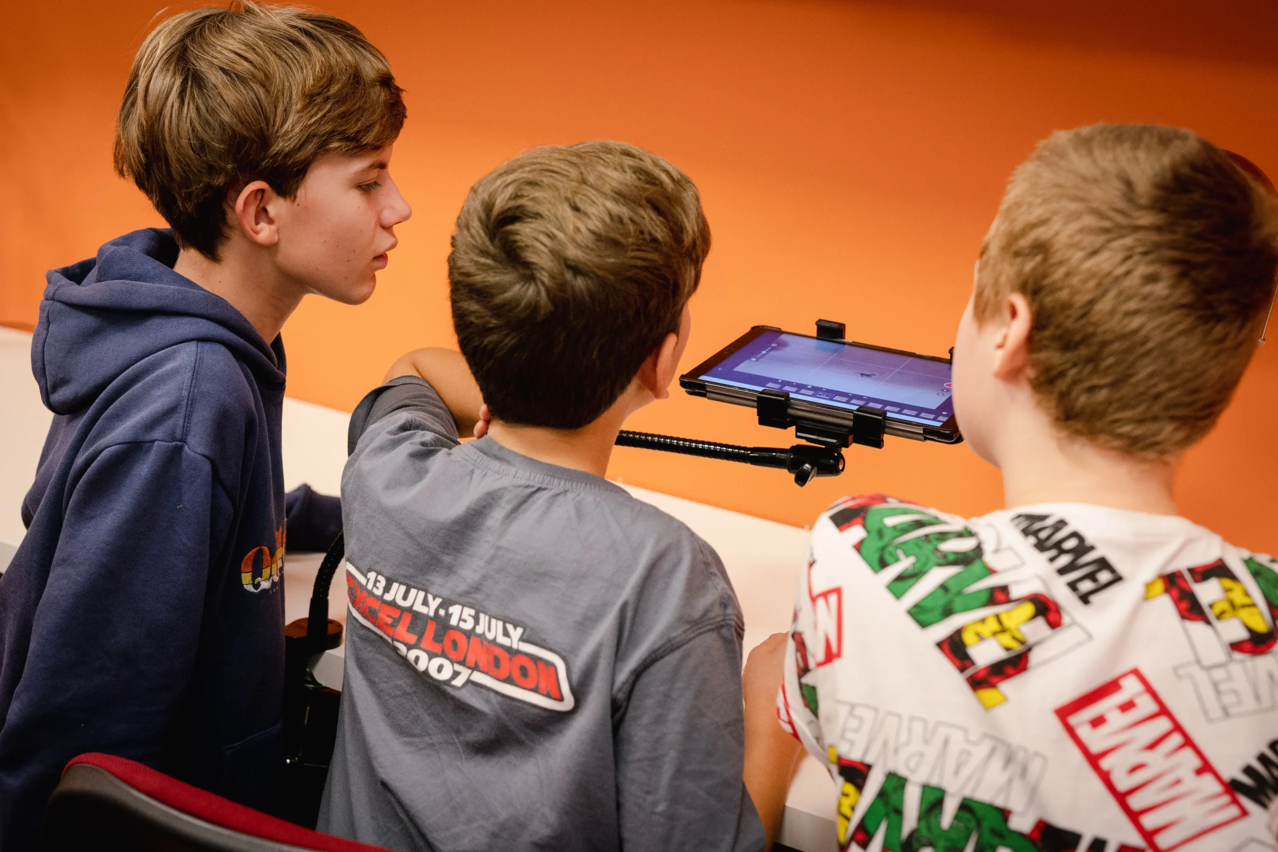 Trzej chłopcy w skupieniu obserwują ekran tabletu zamocowanego na statywie podczas zajęć warsztatowych.