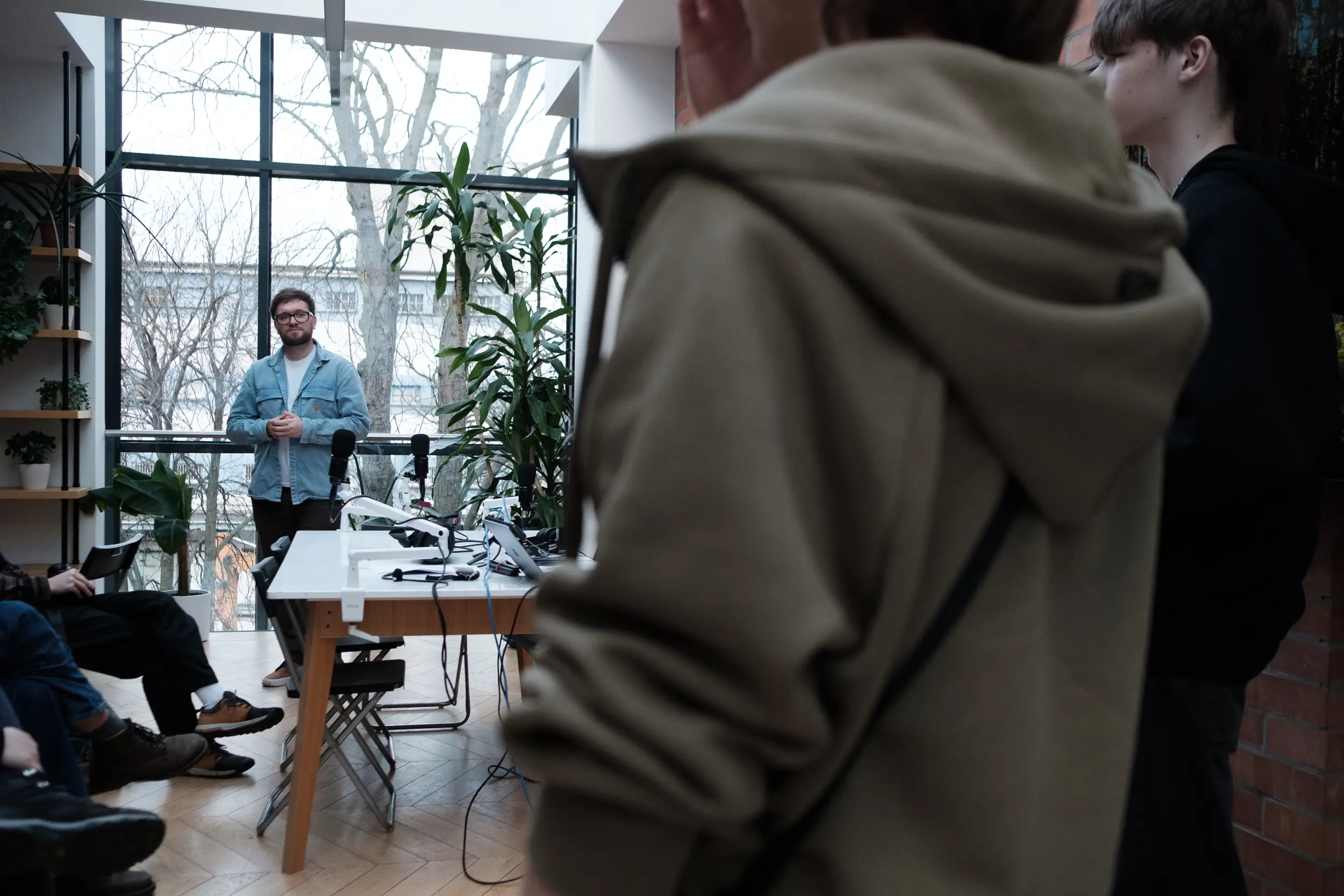 Prowadzący warsztaty podcastowe (Łukasz Laskowski) stoi przy stole ze sprzętem nagraniowym w przeszklonym pomieszczeniu.