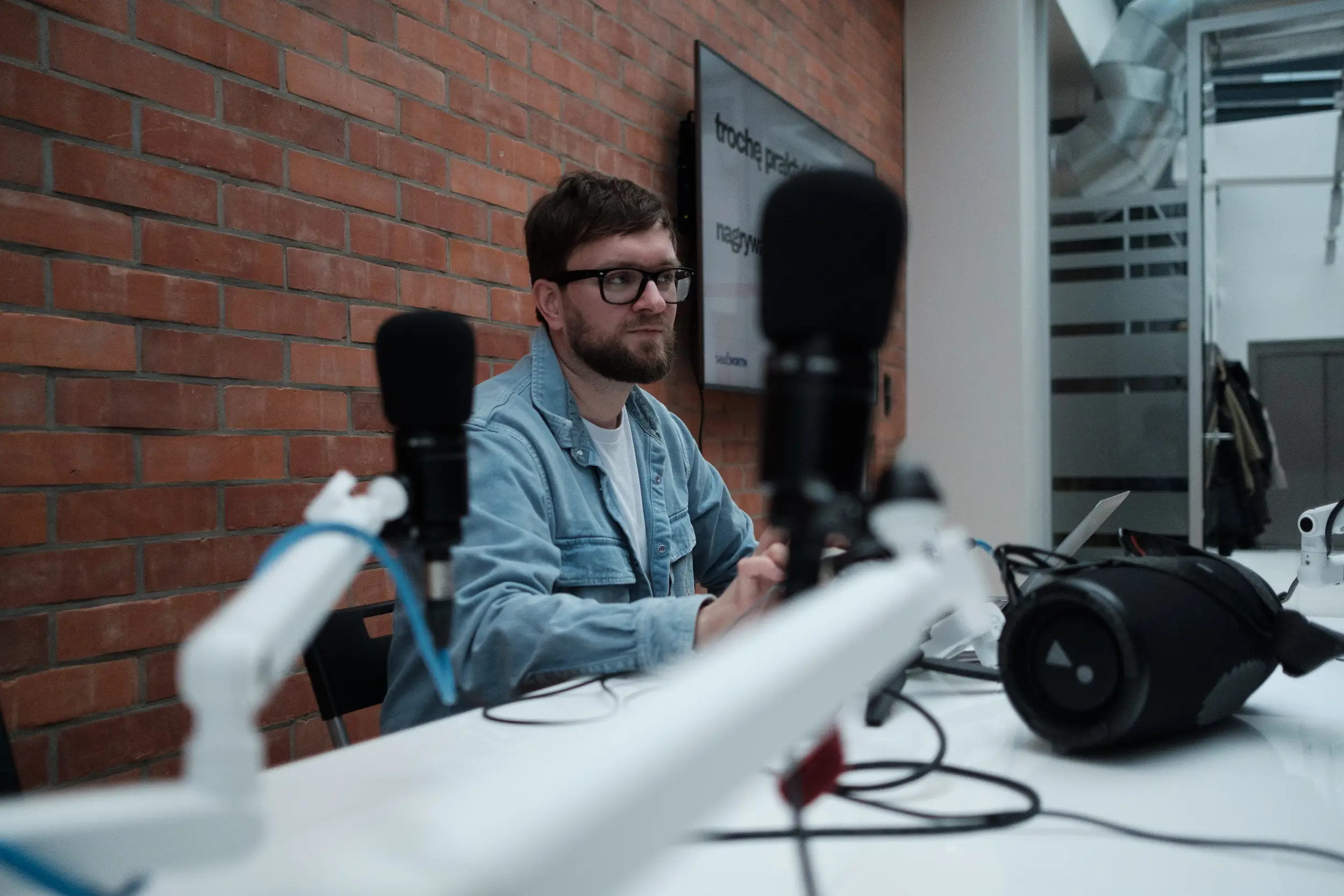 Prowadzący warsztaty w okularach siedzi przy stole z mikrofonami na tle ceglanej ściany i ekranu z prezentacją.