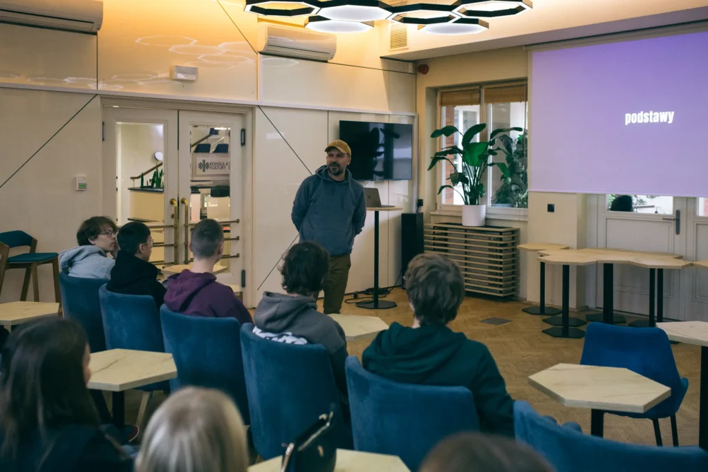Prowadzący w ż&oacute;łtej czapce przemawia do grupy młodzieży siedzącej w niebieskich fotelach podczas warsztat&oacute;w fotograficznych.
