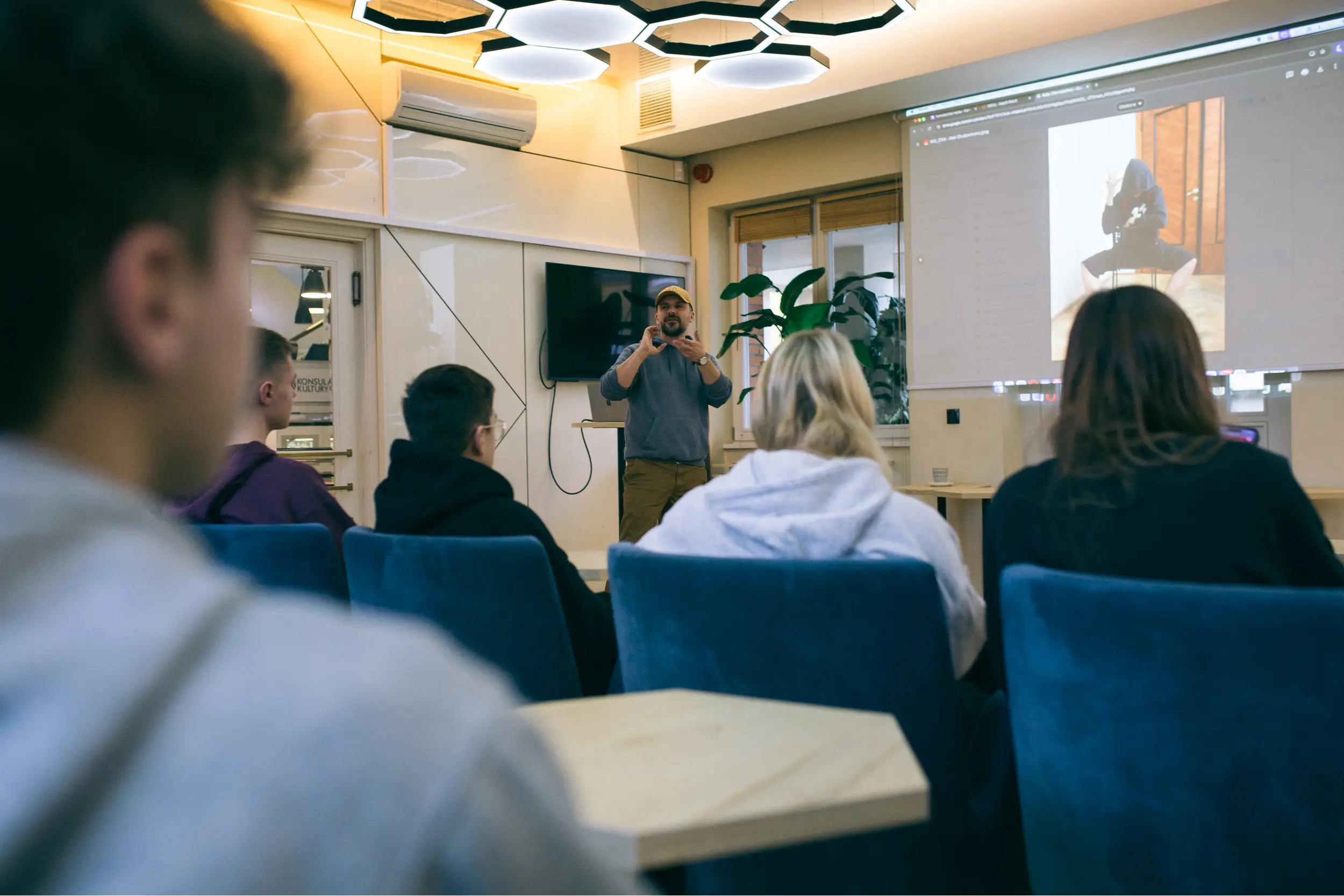 Prowadzący warsztaty gestykuluje na tle dużego ekranu wyświetlającego zdjęcie portretowe przed grupą młodzieży.