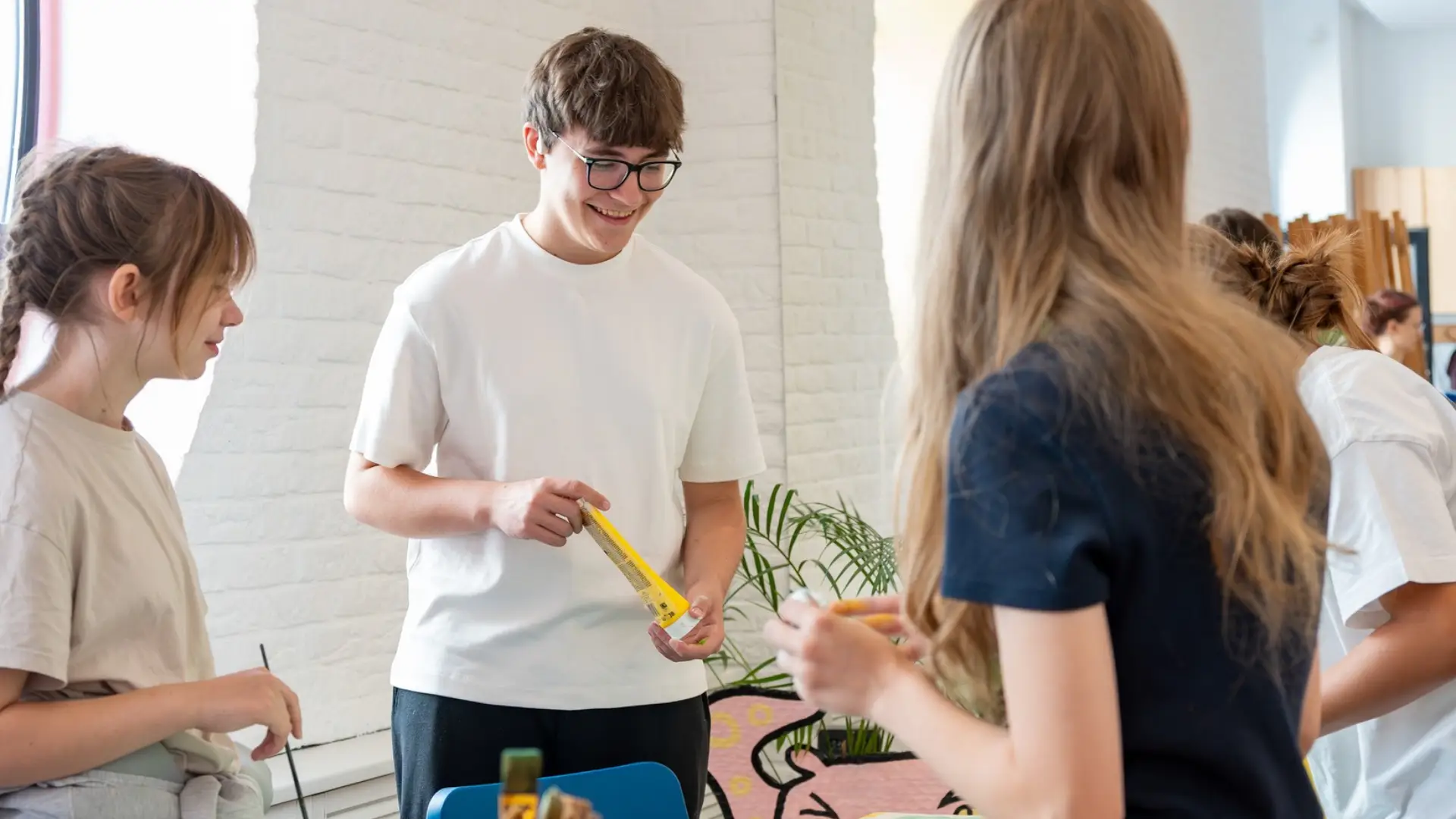 Uśmiechnięta osoba w okularach trzyma żółtą tubkę farby i rozmawia z rówieśnikami podczas warsztatów.