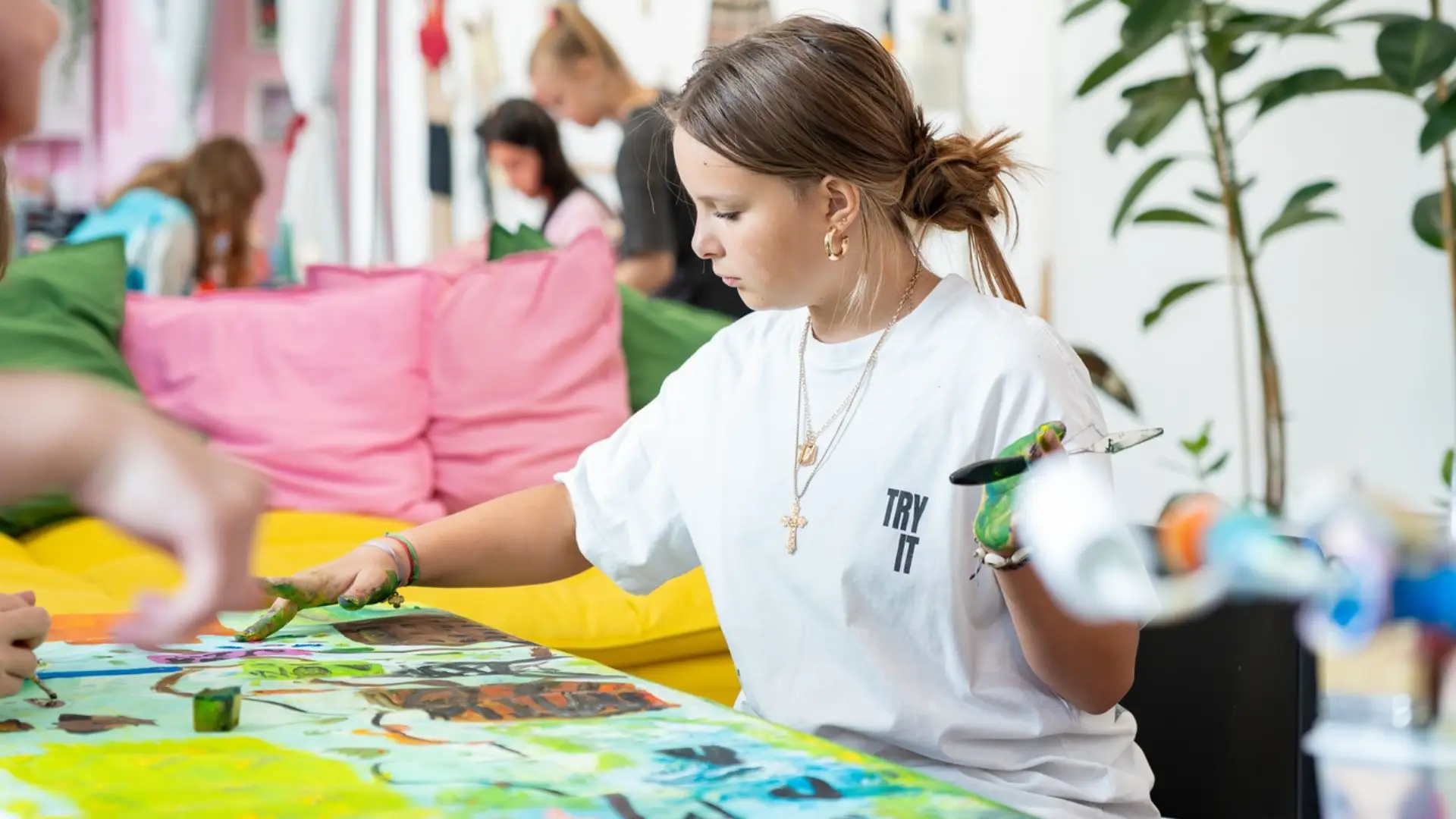 Osoba w białej koszulce z napisem maluje dłonią i szpachelką kolorowy obraz na dużym formacie.