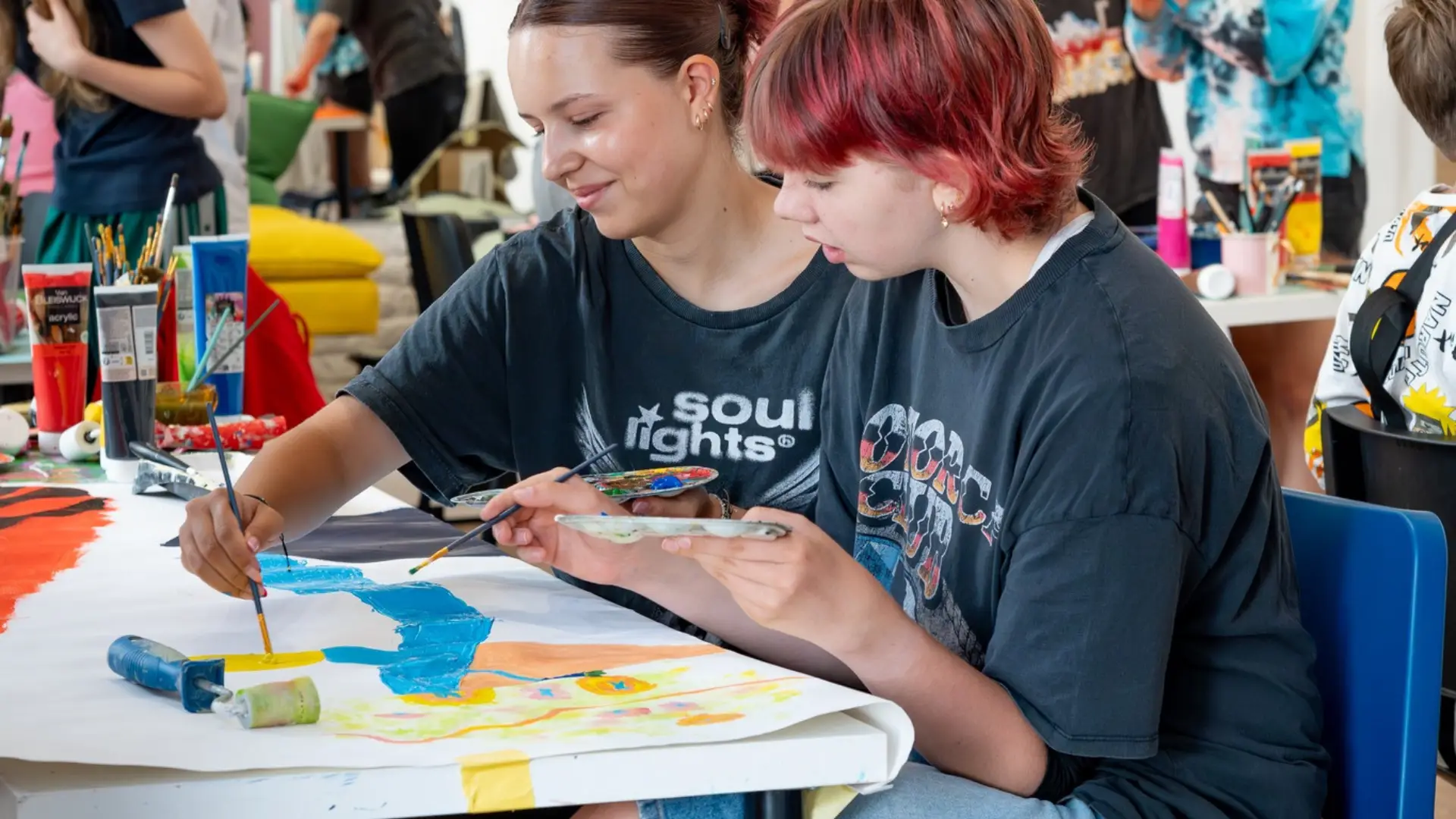 Dwie osoby wspólnie malują pędzlami duży arkusz papieru używając niebieskiej i żółtej farby.