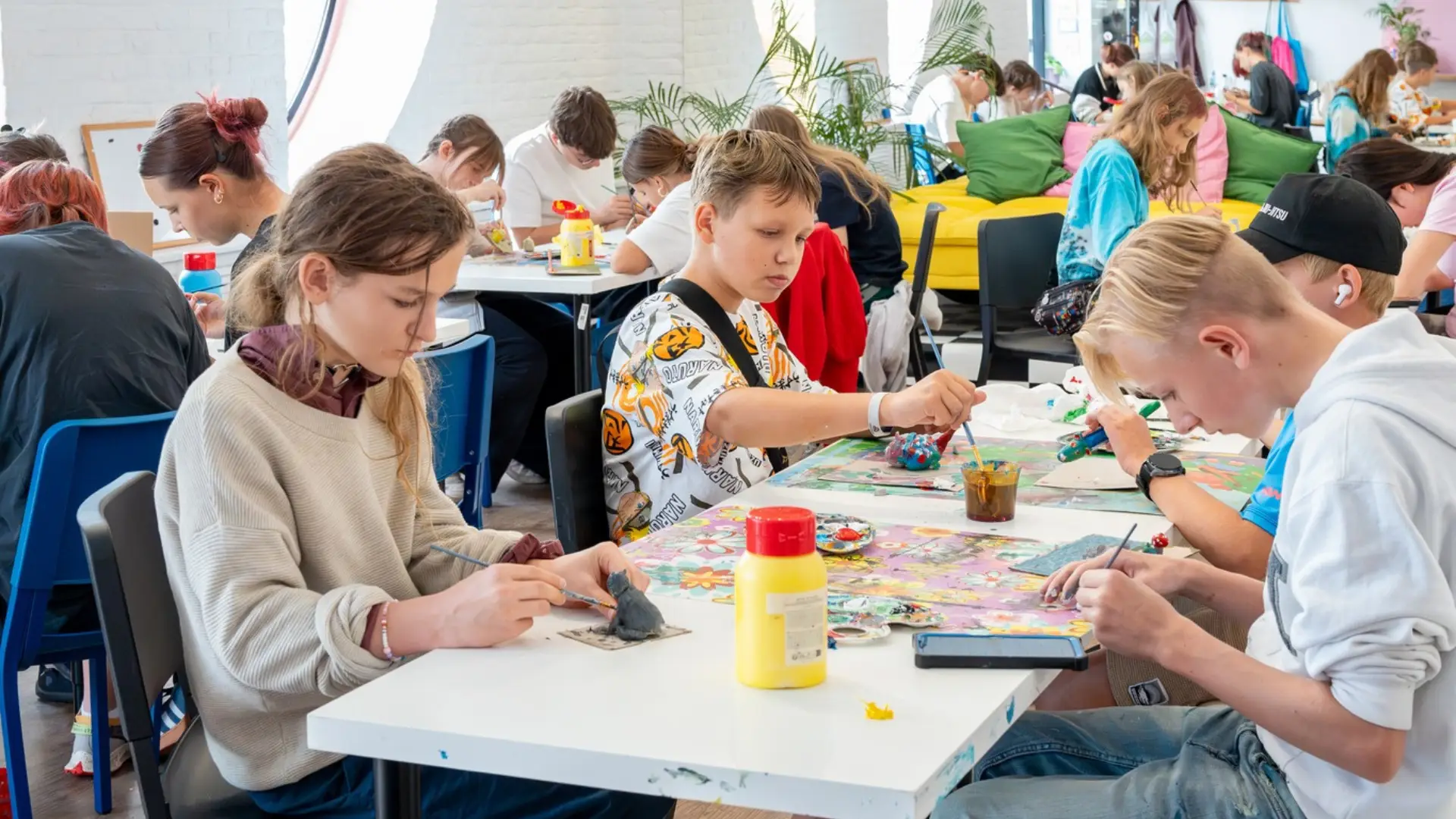 Młodzież skupiona na malowaniu pędzlami małych figurek i form rzeźbiarskich przy białych stolikach.