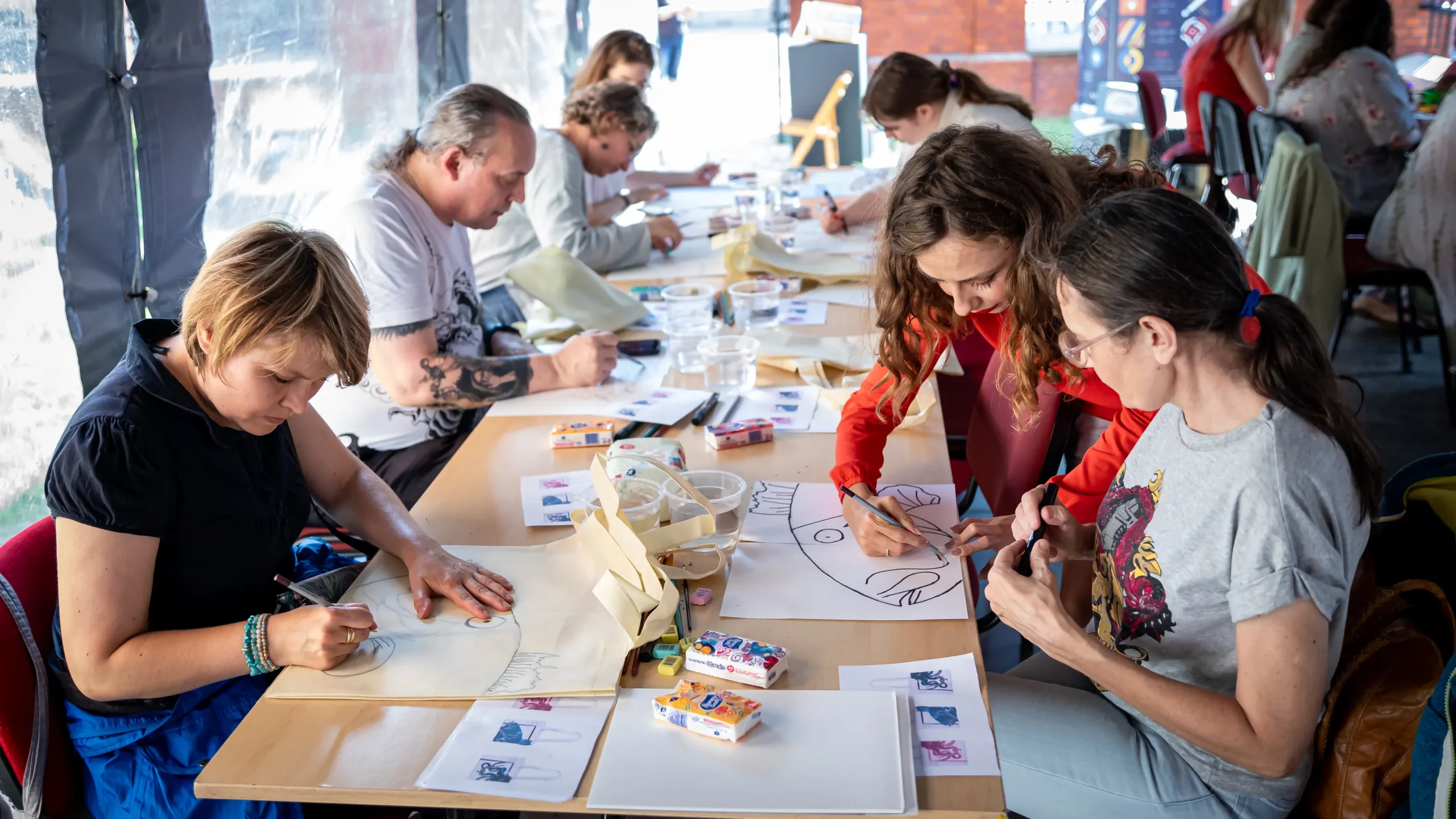 Uczestnicy warsztatów siedzą przy długim stole i nanoszą rysunki na papier oraz torby płócienne.