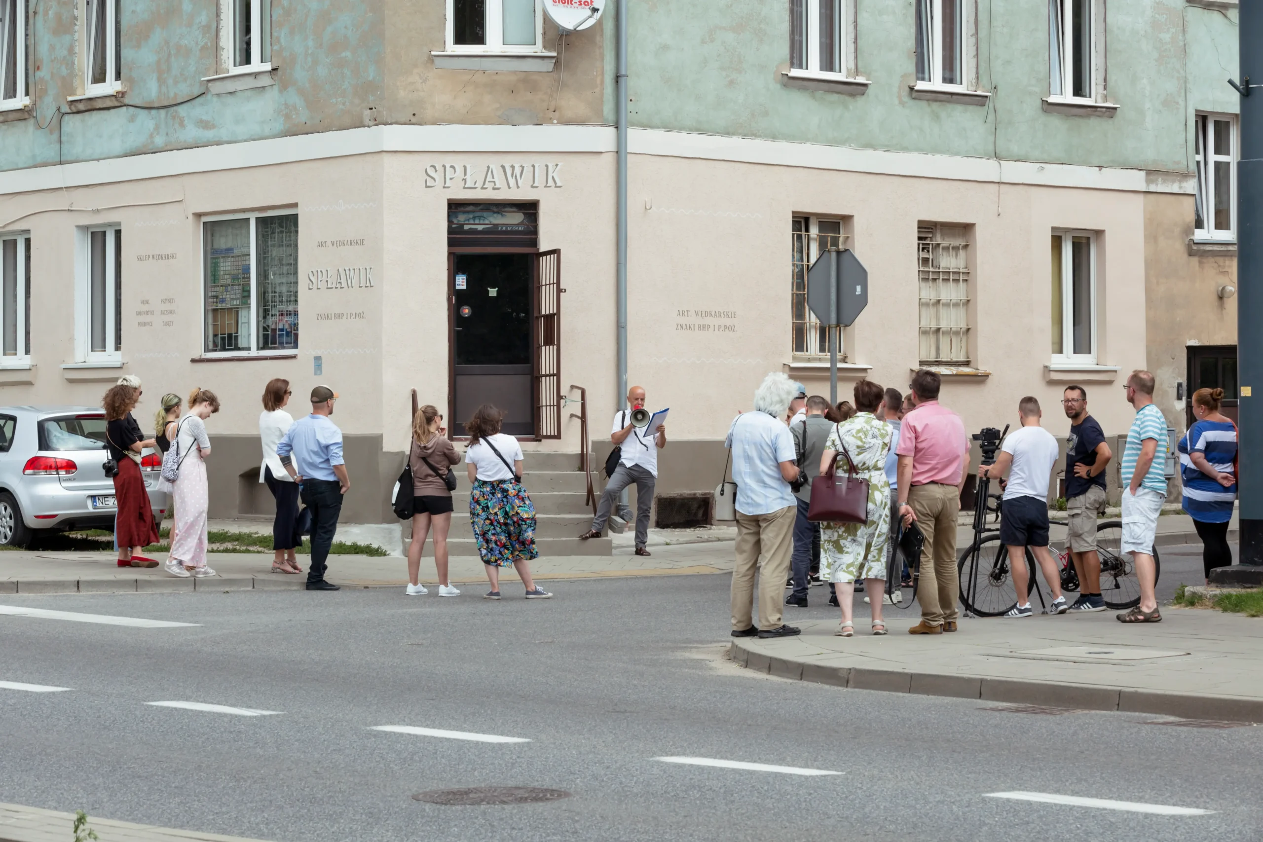 Prowadzący z megafonem i notatkami przemawia do grupy osób stojących na chodniku przed kamienicą ze sklepem wędkarskim.