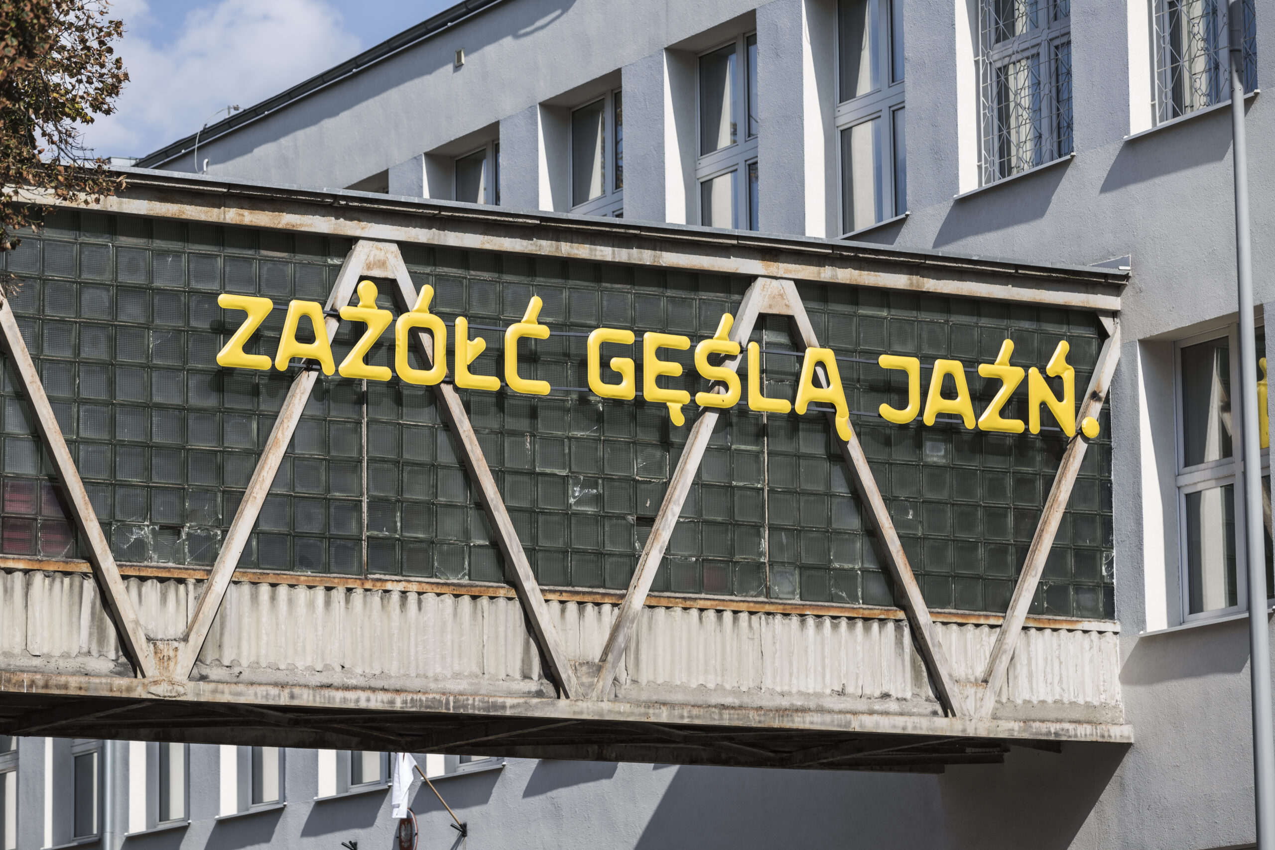 Żółty, trójwymiarowy napis „ZAŻÓŁĆ GĘŚLĄ JAŹŃ” umieszczony na oszklonym łączniku między dwoma budynkami.
