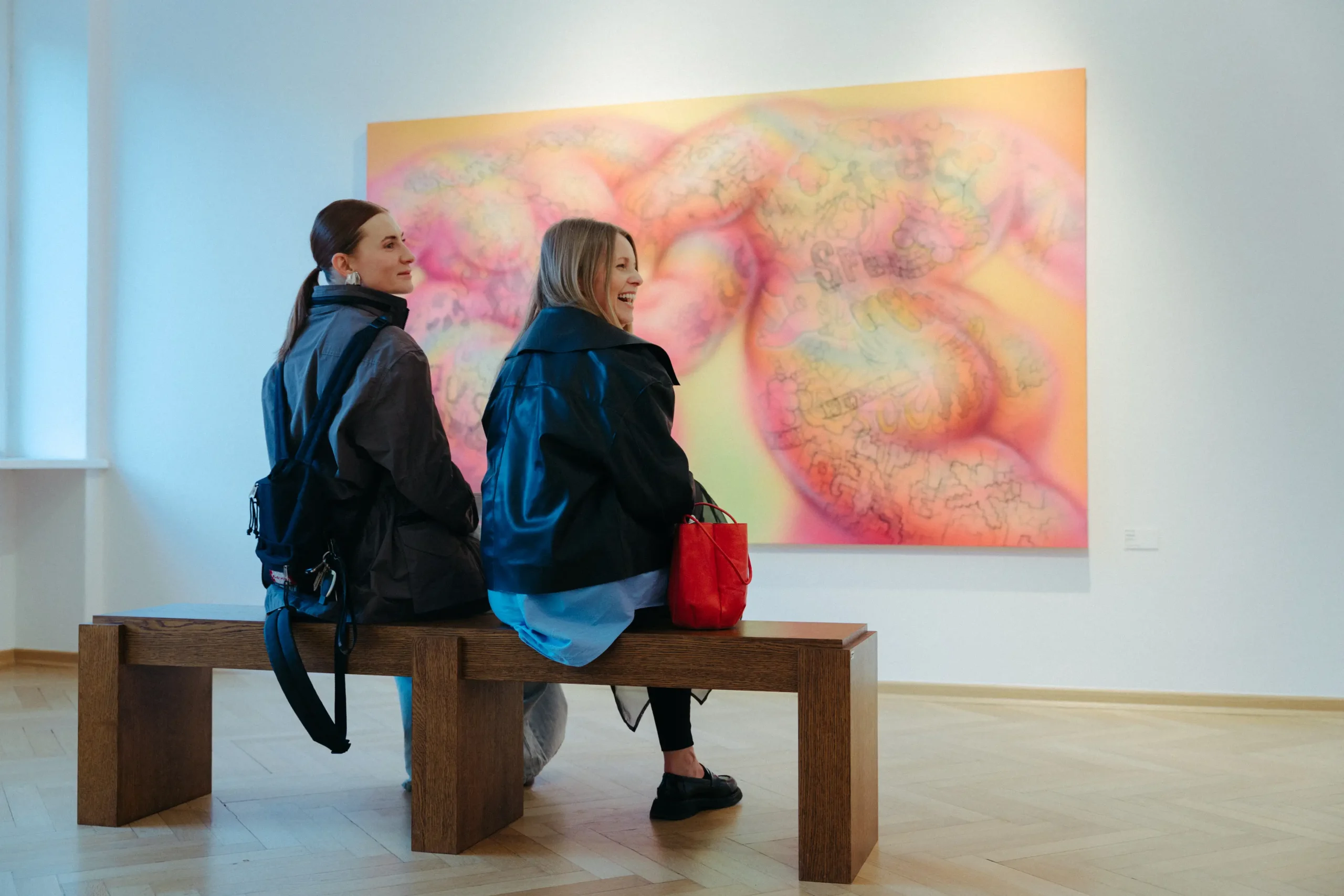 Dwie uśmiechnięte osoby siedzą na drewnianej ławie i oglądają wielkoformatowy obraz w galerii.